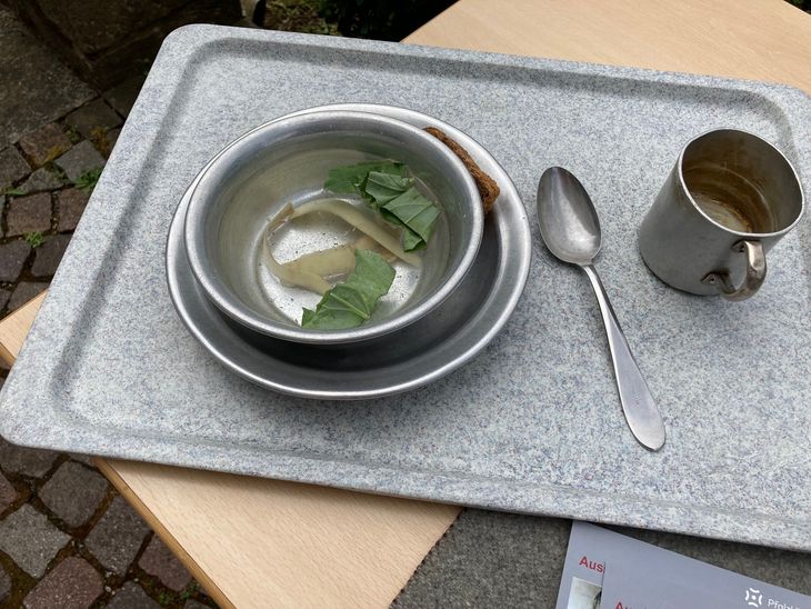 Ein Tablett mit einem Teller und Besteck. Der Teller enthält eine Wassersuppe mit ein paar Gemüseresten.