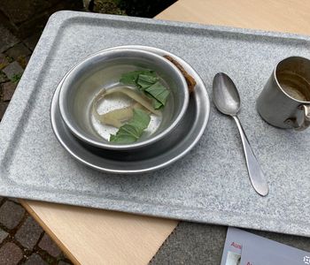 Ein Tablett mit einem Teller und Besteck. Der Teller enthält eine Wassersuppe mit ein paar Gemüseresten.