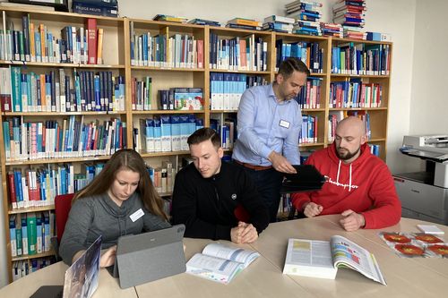 Drei Schüler*innen und eine Lehrkraft in einer Gruppenarbeit vor einem vollen Bücherregal mit medizinischen Fachbüchern
